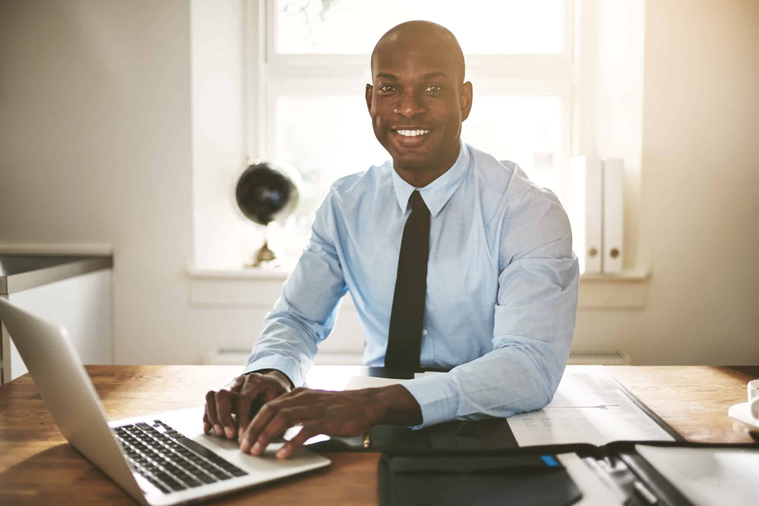 Successful young African businessman sitting at his desk in an office smiling and working on a laptop