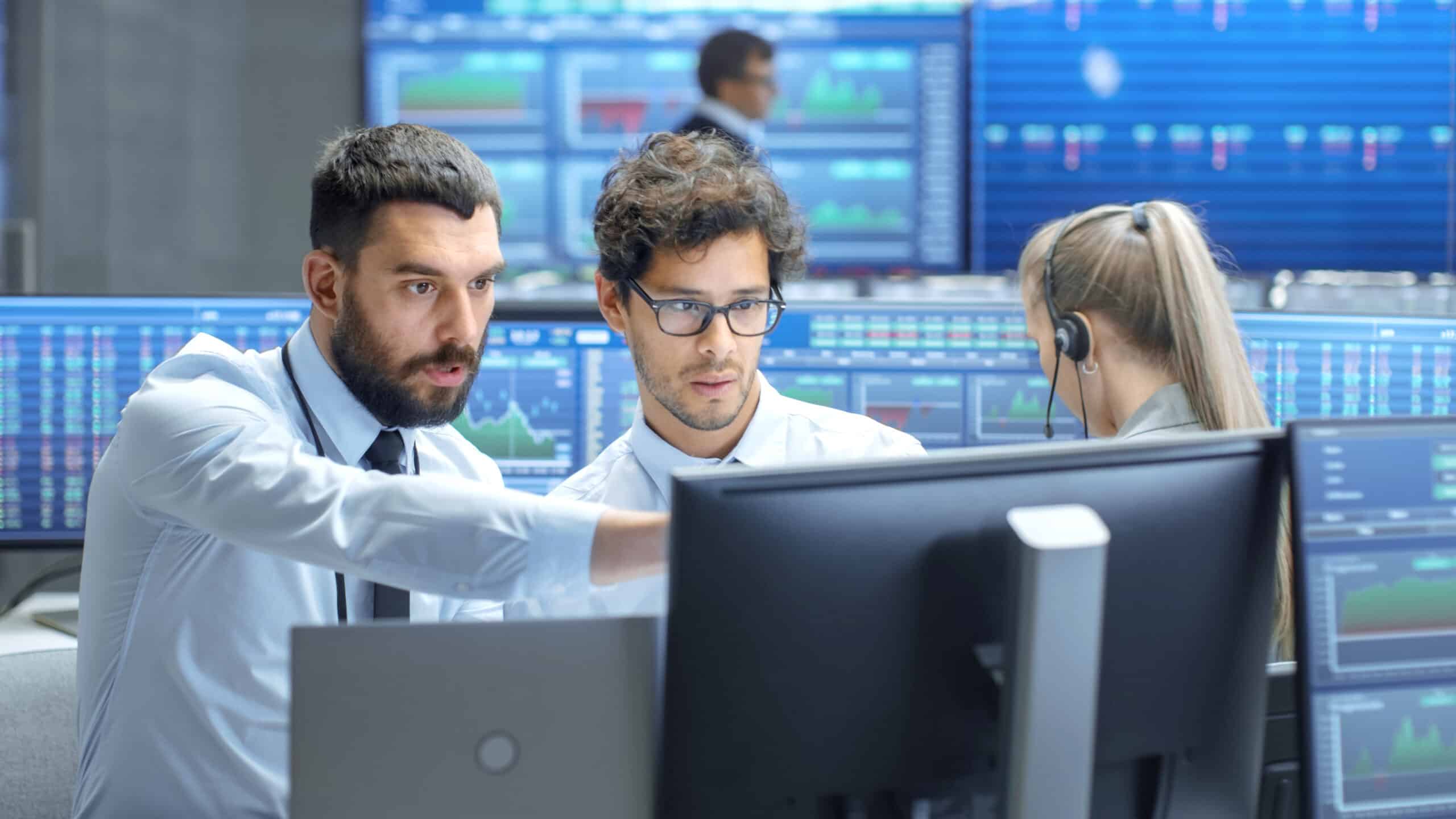 Professional Broker Consults Stock Exchange Trader at His Workstation. Multi-Ethnic Team at Stock Exchange Office is Busy Selling and Buying Stocks on the Market. Displays Show Relevant Data Numbers.